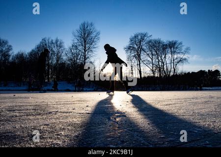 Dunkle Silhouetten von Menschen beim Eislaufen auf dem kristallklaren natürlichen gefrorenen See in der Region Nordbrabant am Nachmittag vor dem Sonnenuntergang. Eislaufrausch übernimmt die Niederlande. Die Niederländer haben viel Spaß und genießen das einwöchige, kalte Schneewetter mit Temperaturen unter Null, die dazu führen, dass Seen, Bäche, Teiche, Flüsse und Kanäle gefrieren. Im Park Meerland in der Nähe der Stadt Eindhoven haben sich die Massen an einem sonnigen Tag mit blauem Himmel abgesetzt, da es eine niederländische Tradition ist. Außerdem werden die Menschen im Freien in der eisgefrorenen Natur und der kalten Umgebung gesehen, die auf dem Eis der gefrorenen Seen spazieren gehen, p Stockfoto