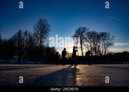 Dunkle Silhouetten von Menschen beim Eislaufen auf dem kristallklaren natürlichen gefrorenen See in der Region Nordbrabant am Nachmittag vor dem Sonnenuntergang. Eislaufrausch übernimmt die Niederlande. Die Niederländer haben viel Spaß und genießen das einwöchige, kalte Schneewetter mit Temperaturen unter Null, die dazu führen, dass Seen, Bäche, Teiche, Flüsse und Kanäle gefrieren. Im Park Meerland in der Nähe der Stadt Eindhoven haben sich die Massen an einem sonnigen Tag mit blauem Himmel abgesetzt, da es eine niederländische Tradition ist. Außerdem werden die Menschen im Freien in der eisgefrorenen Natur und der kalten Umgebung gesehen, die auf dem Eis der gefrorenen Seen spazieren gehen, p Stockfoto
