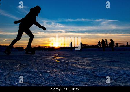 Dunkle Silhouetten von Menschen beim Eislaufen auf dem kristallklaren natürlichen gefrorenen See in der Region Nordbrabant am Nachmittag vor dem Sonnenuntergang. Eislaufrausch übernimmt die Niederlande. Die Niederländer haben viel Spaß und genießen das einwöchige, kalte Schneewetter mit Temperaturen unter Null, die dazu führen, dass Seen, Bäche, Teiche, Flüsse und Kanäle gefrieren. Im Park Meerland in der Nähe der Stadt Eindhoven haben sich die Massen an einem sonnigen Tag mit blauem Himmel abgesetzt, da es eine niederländische Tradition ist. Außerdem werden die Menschen im Freien in der eisgefrorenen Natur und der kalten Umgebung gesehen, die auf dem Eis der gefrorenen Seen spazieren gehen, p Stockfoto