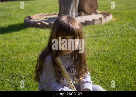 Bild eines entzückenden lächelnden kleinen Mädchens, das auf einem Rasen sitzt und eine Flöte hält. Pädagogische Spiele für Kinder im Freien zu spielen Stockfoto