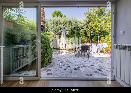 Große Fenster aus weißem Aluminium und Glas öffnen sich in einem Wohnzimmer und bieten Zugang zu einer Terrasse mit Zement- und Steinböden sowie Hecken und Bäumen Stockfoto