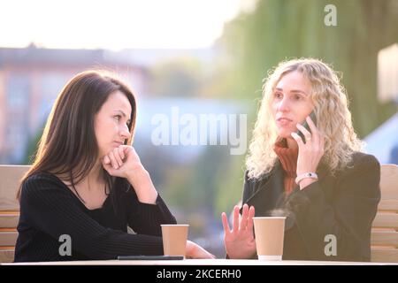 Glückliche junge Frau, die auf dem Handy chattet und ihre gelangweilte Freundin ignoriert. Weibliche Freunde sitzen im Straßencafe im Freien und haben harte Zeit Stockfoto