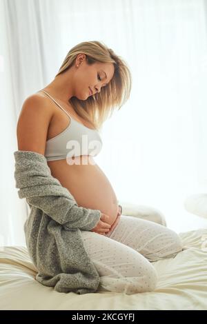 Jede Reise von Müttern ist etwas Besonderes und Unikat. Eine Schwangere berührt ihren Bauch, während sie auf ihrem Bett sitzt. Stockfoto