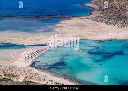 Panoramablick auf den Balos Beach mit den Sonnenschirmen, Sonnendecks und schwimmenden Menschen befindet sich die unglaubliche Lagune mit dem türkisfarbenen exotischen und tropischen Wasser des Mittelmeers in der Region Chania auf der Insel Kreta. Balos ist einer der meistbesuchten Strände Kretas und beliebt bei Besuchern auf der ganzen Welt. Kristallklares Wasser, die Lagune, felsige steile Berge, eine Strandbar mit Sonnenschirmen und Schatten mit Getränken und eine Pirateninsel befinden sich in derselben Region, die mit einem 20-minütigen Trek oder Boot erreichbar ist. Griechenland versucht, seinen Tourismus anzukurbeln und Impfprivilegien zu gewähren Stockfoto
