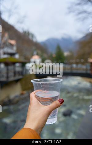 Blick aus der ersten Person mit einem Glas reinem Wasser aus einer Bergquelle vor dem Hintergrund der Stadt Borjomi. Stockfoto