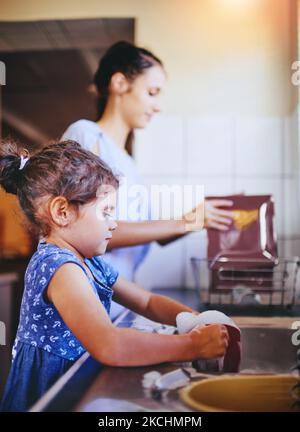 Mummys kleine Helferin. Ein glückliches kleines Mädchen und ihre Mutter waschen zu Hause gemeinsam das Geschirr. Stockfoto