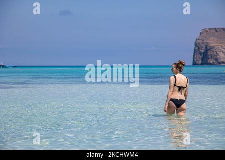 Eine junge Frau, die einen Bikini trägt, wie sie mit ihrer Reflektion auf dem ruhigen Wasser des Balos Beach, der unglaublichen Lagune, mit dem Pool wie türkisblauem, exotischem und tropischem Wasser des Mittelmeers, zu sehen ist, befindet sich in der Region Chania auf der Insel Kreta. Balos ist einer der meistbesuchten Strände Kretas und beliebt bei Besuchern auf der ganzen Welt. Kristallklares Wasser, die Lagune, felsige steile Berge, eine Strandbar mit Sonnenschirmen und Schatten mit Getränken und eine Pirateninsel befinden sich in derselben Region, die mit einem 20-minütigen Trek oder Boot erreichbar ist. Griechenland versucht, seinen Tourismus anzukurbeln und Privilegien zu gewähren Stockfoto