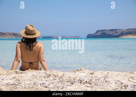 Eine junge Frau mit einem Hut sitzt auf dem weißen rosa Sand des Balos Beach, der unglaublichen Lagune, mit dem Pool wie türkisblauem, exotischem und tropischem Wasser des Mittelmeers in der Region Chania auf der Insel Kreta. Balos ist einer der meistbesuchten Strände Kretas und beliebt bei Besuchern auf der ganzen Welt. Kristallklares Wasser, die Lagune, felsige steile Berge, eine Strandbar mit Sonnenschirmen und Schatten mit Getränken und eine Pirateninsel befinden sich in derselben Region, die mit einem 20-minütigen Trek oder Boot erreichbar ist. Griechenland versucht, seinen Tourismus anzukurbeln und Impfprivilegien zu gewähren Stockfoto