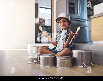 Star, hier komme ich. Porträt eines glücklichen kleinen Jungen, der mit Töpfen auf dem Küchenboden Trommeln spielt, während er eine Schüssel auf dem Kopf trägt. Stockfoto