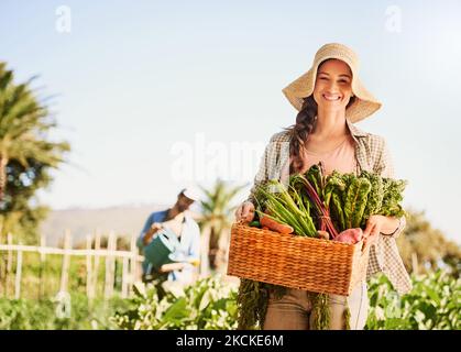 Waren stolz auf unsere Produkte. Porträt einer glücklichen jungen Farmerin, die Kräuter und Gemüse in einem Korb auf ihrem Bauernhof erntet. Stockfoto