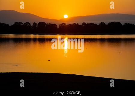 Die Sonne taucht hinter den Bergen auf und reflektiert sich auf der Wasseroberfläche. Magische Stunde Sommer Sonnenaufgang mit warmen Farben des Himmels und der Sonne über Kerkini mit Vögeln, Pferden, lokalen Fischern als Silhouetten gefischt, am See Nationalpark in Serres Region in Nordgriechenland. Der künstliche Kerkini-See ist ein einzigartiges Feuchtgebiet, ein Nationalpark und durch die Ramsar-Konvention als Feuchtgebiet mit Tausenden von Vögeln geschützt, darunter seltene und geschützte Flusswälder, da sich wichtige Hydrobiosphären entwickeln, die von großer internationaler Bedeutung und Akzeptanz sind, die wichtigste Wasserquelle Stockfoto