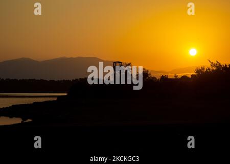 Ein Wachturm für Vogelbeobachter und Besucher des Sees mit der Sonne, die gerade über dem Horizont erscheint. Magische Stunde Sommer Sonnenaufgang mit warmen Farben des Himmels und der Sonne über Kerkini mit Vögeln, Pferden, lokalen Fischern als Silhouetten gefischt, am See Nationalpark in Serres Region in Nordgriechenland. Der künstliche Kerkini-See ist ein einzigartiges Feuchtgebiet, ein Nationalpark und durch die Ramsar-Konvention als Feuchtgebiet mit Tausenden von Vögeln geschützt, darunter seltene und geschützte Wälder am Flussufer, da sich wichtige Hydrobiosphären entwickeln, die von großer internationaler Bedeutung und Akzeptanz sind, t Stockfoto