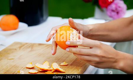 Sommer, im Garten, Nahaufnahme, weibliche Hände schälen eine Orange von der Schale. Familienurlaub, Mittagessen in der Natur, im Hof. Hochwertige Fotos Stockfoto
