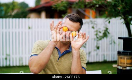 Sommer, im Garten. Ein Mann, Vater, Spaß mit seiner Familie im Freien, Verdrehen, Anwendung Mandarinen auf die Augen. Familienurlaub, Mittagessen in der Natur, im Hof. Hochwertige Fotos Stockfoto