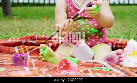 Ein kleines Kind, ein einjähriges Mädchen, das spielt, mit Fingerfarben malt, sich im Garten schmückt, im Sommer auf einer Decke sitzt, auf dem Bettüberwurf, auf Gras, auf dem Rasen. Sie hat Spaß. Hochwertige Fotos Stockfoto