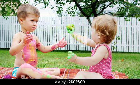Kleine Kinder, ein vierjähriger Junge und ein einjähriges Mädchen, Bruder und Schwester spielen zusammen, malen mit Fingerfarben, im Garten, sitzen auf einer Decke, auf Gras, Rasen, im Sommer. Hochwertige Fotos Stockfoto
