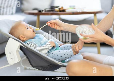 Mutter Löffel Fütterung ihres kleinen Jungen Kind in Babystuhl mit Fruchtpüree. Baby feste Nahrung Einführung Konzept Stockfoto