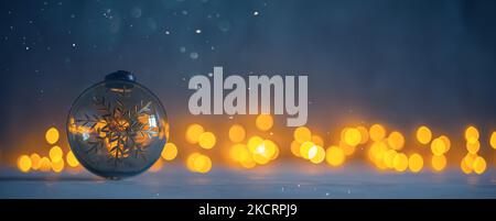 Weihnachtsbaumkugel aus Glas auf dunklem Hintergrund mit Schnee und glühenden goldenen Bokeh-Lichtern Stockfoto