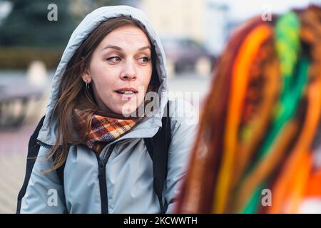 Junge Frauen sprechen, sitzen im Stadtpark. Zwei Freundinnen in warmen Kleidern plaudern auf der Straße. Selektiver Fokus auf emotionale Frauen Stockfoto