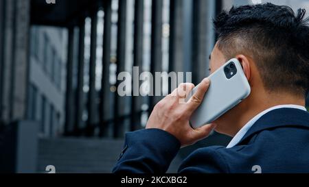Rückansicht von hinten nicht erkennbarer asiatischer Mann in formellem Anzug, der in der Stadt steht und mit dem Smartphone telefoniert und den Chef anruft Stockfoto