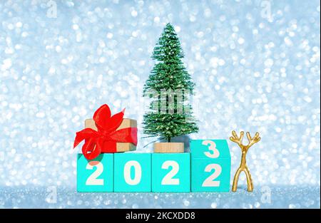 Jahr 2022 bis 2023 Übergangshintergrund mit Holzblöcken, Spielzeug-weihnachtsbaum, winziger Hirschfigur und dekorierter Geschenkbox vor einem glitzernden Backdro Stockfoto