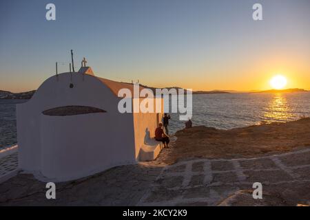 Sonnenuntergang mit einer kleinen, weiß getünchten Kirche, einer Kapelle mit Menschen, die den Sonnenuntergang fotografieren. Touristen genießen einen wunderschönen Sonnenuntergang während der magischen Stunde auf der Insel Mykonos in Griechenland. Die griechische Insel Myconos ist ein beliebtes glamouröses mediterranes Reiseziel für einen Urlaub in den Kykladen, der Ägäis mit den ikonischen weiß getünchten Gebäuden, den Sandstränden und berühmten Partys an den Strandbars. Die Tourismus- und Reisebranche hatte aufgrund der Coronavirus-Pandemie Covid-19 negative Auswirkungen auf die Wirtschaft und die lokale Wirtschaft. Mykonos, Griechenland am 10. Oktober 2021 (Foto: Nicolas E Stockfoto