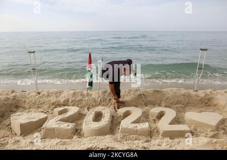 Ein Palästinenser Mohammed Totah ''32'', der am Strand von Gaza eine Sandzeichnung von 2022 anstellt und seine Hoffnung zum Ausdruck bringt, dass das neue Jahr das Friedensjahr Palästinas sein wird, am 28. Dezember 2021. Mohammed wurde nach israelischen Angriffen während des Gaza-Krieges am 28. Dezember 2008 im Führungskrieg gegen den Gazastreifen verletzt, der sein rechtes Bein verlor. Mohammed spricht über seine verheerende Verletzung, die sein Leben veränderte, ihn aber nicht von seinem Traum und seinem Streben nach einem guten Leben mit seiner Familie abgehalten hat. (Foto von Sameh Rahmi/NurPhoto) Stockfoto