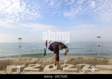 Ein Palästinenser Mohammed Totah ''32'', der am Strand von Gaza eine Sandzeichnung von 2022 anstellt und seine Hoffnung zum Ausdruck bringt, dass das neue Jahr das Friedensjahr Palästinas sein wird, am 28. Dezember 2021. Mohammed wurde nach israelischen Angriffen während des Gaza-Krieges am 28. Dezember 2008 im Führungskrieg gegen den Gazastreifen verletzt, der sein rechtes Bein verlor. Mohammed spricht über seine verheerende Verletzung, die sein Leben veränderte, ihn aber nicht von seinem Traum und seinem Streben nach einem guten Leben mit seiner Familie abgehalten hat. (Foto von Sameh Rahmi/NurPhoto) Stockfoto