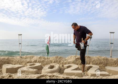 Ein Palästinenser Mohammed Totah ''32'', der am Strand von Gaza eine Sandzeichnung von 2022 anstellt und seine Hoffnung zum Ausdruck bringt, dass das neue Jahr das Friedensjahr Palästinas sein wird, am 28. Dezember 2021. Mohammed wurde nach israelischen Angriffen während des Gaza-Krieges am 28. Dezember 2008 im Führungskrieg gegen den Gazastreifen verletzt, der sein rechtes Bein verlor. Mohammed spricht über seine verheerende Verletzung, die sein Leben veränderte, ihn aber nicht von seinem Traum und seinem Streben nach einem guten Leben mit seiner Familie abgehalten hat. (Foto von Sameh Rahmi/NurPhoto) Stockfoto