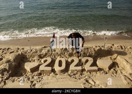 Ein Palästinenser Mohammed Totah ''32'', der am Strand von Gaza eine Sandzeichnung von 2022 anstellt und seine Hoffnung zum Ausdruck bringt, dass das neue Jahr das Friedensjahr Palästinas sein wird, am 28. Dezember 2021. Mohammed wurde nach israelischen Angriffen während des Gaza-Krieges am 28. Dezember 2008 im Führungskrieg gegen den Gazastreifen verletzt, der sein rechtes Bein verlor. Mohammed spricht über seine verheerende Verletzung, die sein Leben veränderte, ihn aber nicht von seinem Traum und seinem Streben nach einem guten Leben mit seiner Familie abgehalten hat. (Foto von Sameh Rahmi/NurPhoto) Stockfoto