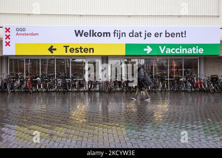 Eine Frau, die unter einem Banner mit Anweisungen für das Test- oder Impfzentrum in Richtung der Testeinrichtung geht. Menschen mit Gesichtsmasken warten vor einer Einrichtung des GGD Paviljoen Amsterdam Covid-19-Testzentrums, um getestet zu werden, GGD ist der städtische Gesundheitsdienst in Amsterdam Zuid. Die Niederlande waren die erste europäische Nation, die kurz vor Weihnachten während der Coronavirus-Pandemie von Covid-19 eine Sperre gegen die neue Omicron-Variante ausrief. Bars, Fitnessstudios, Friseure und alle „nicht notwendigen“ Geschäfte, Museen, öffentlichen Einrichtungen usw. waren bis mindestens Januar 14 geschlossen. Amste Stockfoto