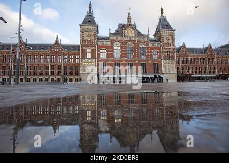 Tägliches Leben mit Menschen, wie man es mit dem Amsterdamer Hauptbahnhof im Hintergrund sieht. Einheimische und ein paar Touristen in den ruhigen Straßen von Amsterdam während der Sperre in der niederländischen Hauptstadt mit Geschäften und Geschäften, die mit geschlossenen Rollläden aus Metall erscheinen, Cafés, Bars und Restaurants auch geschlossen mit Tischen und Stühlen der Terrassen verschlossen. Die Niederlande waren die erste europäische Nation, die die vollständige Sperre erklärte, um gegen die neue Omicron-Variante zu kämpfen, die aufkommt. Nach einem plötzlichen Regierungsbefehl vor Weihnachten schloss das Land alle unwesentlichen Geschäfte, Cafés, Restaurants, Stockfoto