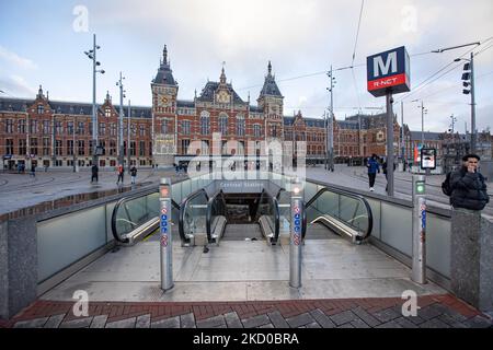 Das tägliche Leben mit den Menschen, wie man es mit dem U-Bahn-Eingang und dem Hauptbahnhof von Amsterdam im Hintergrund sieht. Einheimische und ein paar Touristen in den ruhigen Straßen von Amsterdam während der Sperre in der niederländischen Hauptstadt mit Geschäften und Geschäften, die mit geschlossenen Rollläden aus Metall erscheinen, Cafés, Bars und Restaurants auch geschlossen mit Tischen und Stühlen der Terrassen verschlossen. Die Niederlande waren die erste europäische Nation, die die vollständige Sperre erklärte, um gegen die neue Omicron-Variante zu kämpfen, die aufkommt. Nach einem plötzlichen Regierungsbefehl vor Weihnachten schloss das Land alle Nichtessen Stockfoto