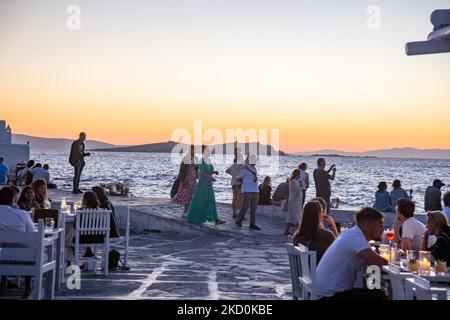 Die Menschen genießen den Sonnenuntergang von Little Venice. Little Venice auf der Insel Mykonos während der magischen Stunde des Sonnenuntergangs. Touristen genießen einen Drink auf der Terrasse oder dem Balkon der weiß getünchten Café-Bars oder ein Abendessen in einem Restaurant direkt über dem Wasser in dem kleinen Hafengebiet unter den berühmten Windmühlen der Insel. Die griechische Insel Myconos ist ein beliebtes glamouröses mediterranes Reiseziel für einen Urlaub in den Kykladen, der Ägäis mit den ikonischen weiß getünchten Gebäuden, den Sandstränden und berühmten Partys an den Strandbars. Die Tourismus- und Reisebranche hatte negative Auswirkungen auf den Geschäftsverkehr Stockfoto