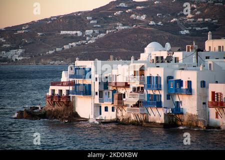 Panoramablick auf Little Venice auf der Insel Mykonos während der magischen Stunde des Sonnenuntergangs. Touristen genießen einen Drink auf der Terrasse oder dem Balkon der weiß getünchten Café-Bars oder ein Abendessen in einem Restaurant direkt über dem Wasser in dem kleinen Hafengebiet unter den berühmten Windmühlen der Insel. Die griechische Insel Myconos ist ein beliebtes glamouröses mediterranes Reiseziel für einen Urlaub in den Kykladen, der Ägäis mit den ikonischen weiß getünchten Gebäuden, den Sandstränden und berühmten Partys an den Strandbars. Die Tourismus- und Reisebranche hatte negative Auswirkungen auf die Wirtschaft und die lokale Wirtschaft Stockfoto