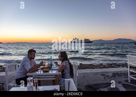 Ein Paar hat Abendessen während des Sonnenuntergangs in Little Venice. Little Venice auf der Insel Mykonos während der magischen Stunde des Sonnenuntergangs. Touristen genießen einen Drink auf der Terrasse oder dem Balkon der weiß getünchten Café-Bars oder ein Abendessen in einem Restaurant direkt über dem Wasser in dem kleinen Hafengebiet unter den berühmten Windmühlen der Insel. Die griechische Insel Myconos ist ein beliebtes glamouröses mediterranes Reiseziel für einen Urlaub in den Kykladen, der Ägäis mit den ikonischen weiß getünchten Gebäuden, den Sandstränden und berühmten Partys an den Strandbars. Die Tourismus- und Reisebranche hatte negative Auswirkungen auf die Stockfoto