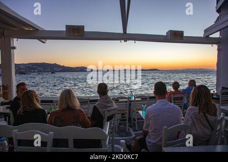 Die Menschen genießen den Sonnenuntergang von Little Venice. Little Venice auf der Insel Mykonos während der magischen Stunde des Sonnenuntergangs. Touristen genießen einen Drink auf der Terrasse oder dem Balkon der weiß getünchten Café-Bars oder ein Abendessen in einem Restaurant direkt über dem Wasser in dem kleinen Hafengebiet unter den berühmten Windmühlen der Insel. Die griechische Insel Myconos ist ein beliebtes glamouröses mediterranes Reiseziel für einen Urlaub in den Kykladen, der Ägäis mit den ikonischen weiß getünchten Gebäuden, den Sandstränden und berühmten Partys an den Strandbars. Die Tourismus- und Reisebranche hatte negative Auswirkungen auf den Geschäftsverkehr Stockfoto