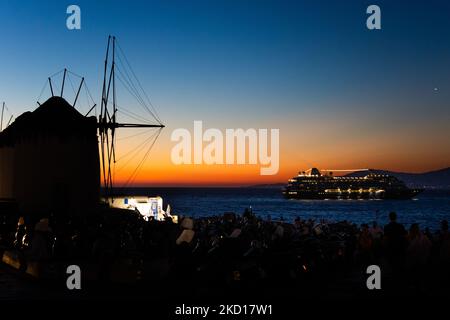 Magische Stunde bei den berühmten Windmühlen der Insel Mykonos mit einem Schiff sichtbar, auf den Kykladen Inseln, Griechenland. Mykonos ist eine Insel in Griechenland in der Ägäis. Auf der Insel gibt es 16 Windmühlen, 5 davon oberhalb von Chora, dem Hauptort der Insel. Die Windmühlen wurden im 16.. Jahrhundert von den Venezianern gebaut, aber ihre Bauten dauerten bis zum 20.. Jahrhundert an. Sonnenuntergang mit Menschen an den Windmühlen auf Mykonos. Touristen genießen einen wunderschönen Sonnenuntergang während der magischen Stunde auf der Insel Mykonos in Griechenland. Die griechische Insel Myconos ist ein beliebtes glamouröses mediterranes Urlaubsziel Stockfoto