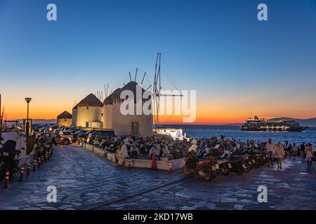 Magische Stunde bei den berühmten Windmühlen der Insel Mykonos mit einem Schiff sichtbar, auf den Kykladen Inseln, Griechenland. Mykonos ist eine Insel in Griechenland in der Ägäis. Auf der Insel gibt es 16 Windmühlen, 5 davon oberhalb von Chora, dem Hauptort der Insel. Die Windmühlen wurden im 16.. Jahrhundert von den Venezianern gebaut, aber ihre Bauten dauerten bis zum 20.. Jahrhundert an. Sonnenuntergang mit Menschen an den Windmühlen auf Mykonos. Touristen genießen einen wunderschönen Sonnenuntergang während der magischen Stunde auf der Insel Mykonos in Griechenland. Die griechische Insel Myconos ist ein beliebtes glamouröses mediterranes Urlaubsziel Stockfoto