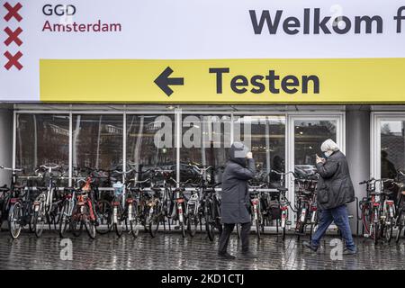 Menschen mit Gesichtsmasken gehen unter ein großes Schild, das die Impfstelle und den Testort zeigt. Tägliches Leben während der Sperre bei der vierten Welle der Pandemie. Einheimische und ein paar Touristen in den ruhigen Straßen von Amsterdam während der Sperre in der niederländischen Hauptstadt mit Geschäften und Geschäften, die mit geschlossenen Rollläden aus Metall erscheinen, Cafés, Bars und Restaurants auch geschlossen mit Tischen und Stühlen der Terrassen verschlossen. Die Niederlande waren die erste europäische Nation, die die vollständige Sperre erklärte, um gegen die neue Omicron-Variante zu kämpfen, die aufkommt. Nach einem plötzlichen Regierungsbefehl vor Ch Stockfoto