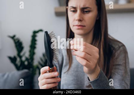 Nahaufnahme einer unglücklich frustrierten, langhaarigen jungen Frau, die viele Haare in ihrer Hand betrachtet und auf der Couch sitzt. Haarausfall, Alopezie, Symptome Stockfoto