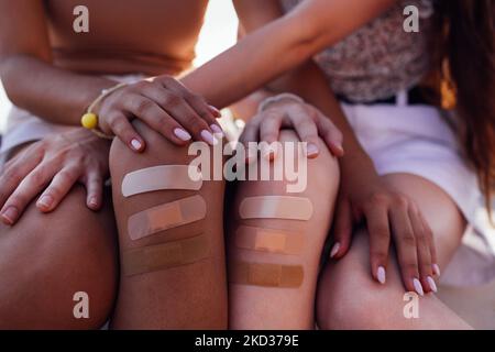 Beschnittene Ansicht von zwei verschiedenen Rassen weibliche Teenager-Freundinnen mit klebenden Pflaster auf verletzten Knie sitzen auf der Bordbank. Diversity Peaple und Care Conce Stockfoto