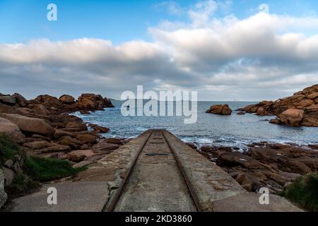 Frankreich, Ploumenach, 2022-01-13. Die Schienen für die Boote laufen entlang der rosafarbenen Granitküste in der Bretagne mit der Spitze der gemeinen Ruz Lig ins Wasser Stockfoto