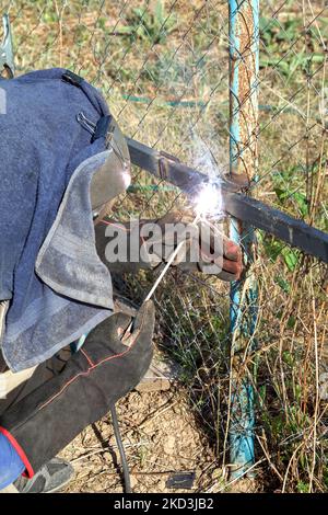 Schweißer in braunem Overall, Schutzmaske und Handschuhe verschweißt Metallrohre an senkrechten Basen für die weitere Befestigung von Metallblechen mit elektrischen ar Stockfoto