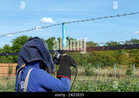 Ein Arbeiter in der Schwerindustrie schleift Schweißnähte mit einem elektrischen Winkelschleifer auf seiner Baustelle. Die Maske und die Schutzhandschuhe schützen vor Funken in den Augen und Stockfoto