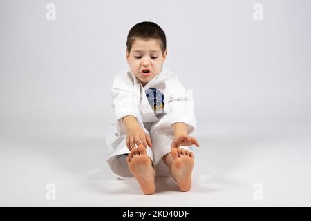 Ein kleiner Junge im Kimono, der Karate macht, streckt seine Hände bis an die Zehenspitzen, übt sich auf weißem Hintergrund zu strecken. Stockfoto