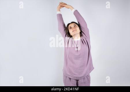 Junge Brünette Frau in Musselin Lavendel Schlafanzug Schlafanzug erstreckt sich, indem sie ihre Arme nach oben. Stockfoto