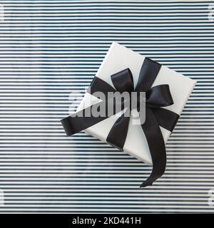 Geburtstag Jubiläum Weihnachten Valentinstag Geschenk. Eine weiße Geschenkschachtel mit luxuriösem schwarzen Satinband auf gestreiftem Hintergrund, Draufsicht. Stockfoto