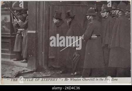 „The Battle of Stepney: Mr. Winston Churchill in the Fire Zone“: Postkarte der Belagerung der Sidney Street von 1911, auf der Innenminister Winston Churchill in einem Zylinderhut die Sidney Street in das belagerte Haus spähend sieht. Soldaten mit Gewehren stehen vor ihm und eine Gruppe von Polizisten hinter ihm. Stockfoto