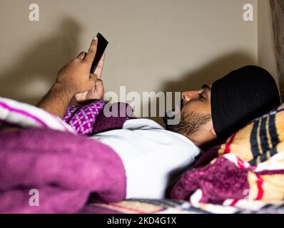 Eine Nahaufnahme eines südasiatischen Mannes, der in einem Bett liegt, mit dem Turban auf dem Kopf, bedeckt mit einer Patchwork-Steppdecke, und auf ein schwarzes Mobiltelefon blickt Stockfoto